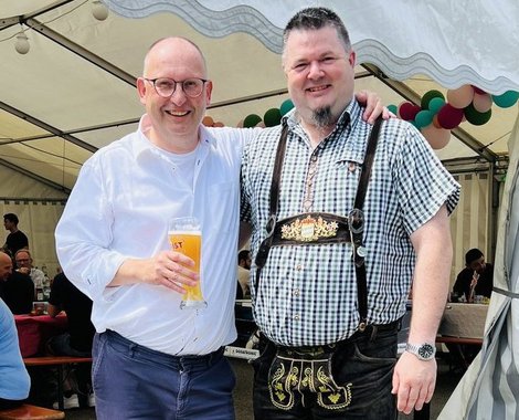 Zwei Männer beim Bierfest unter einem Zelt: einer hält Bierglas, der andere in Trachtenlook; beide lächeln in die Kamera.