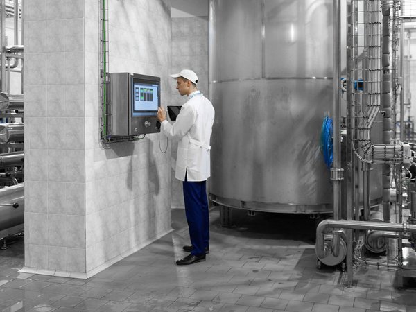 Mitarbeiter im weißen Kittel bedient ein Touchscreen vor einer großen Edelstahlanlage in einer Industriehalle.